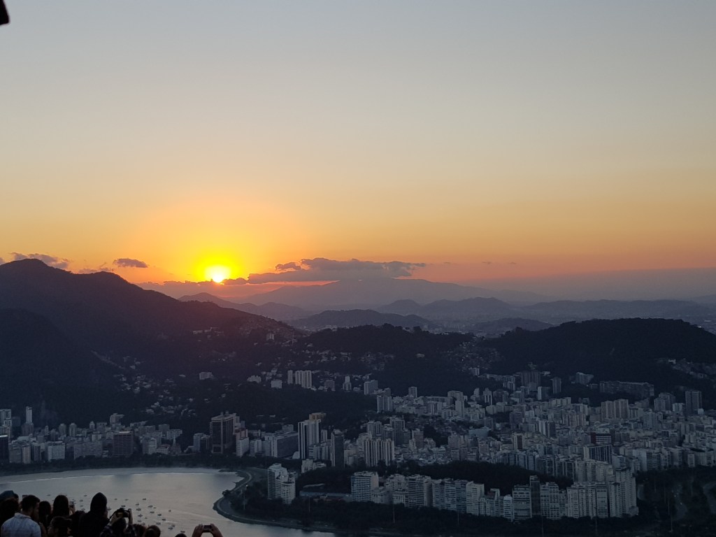 Puesta de sol sobre las montañas y la ciudad desde el Pan de Azúcar, con tonos cálidos en el horizonte.